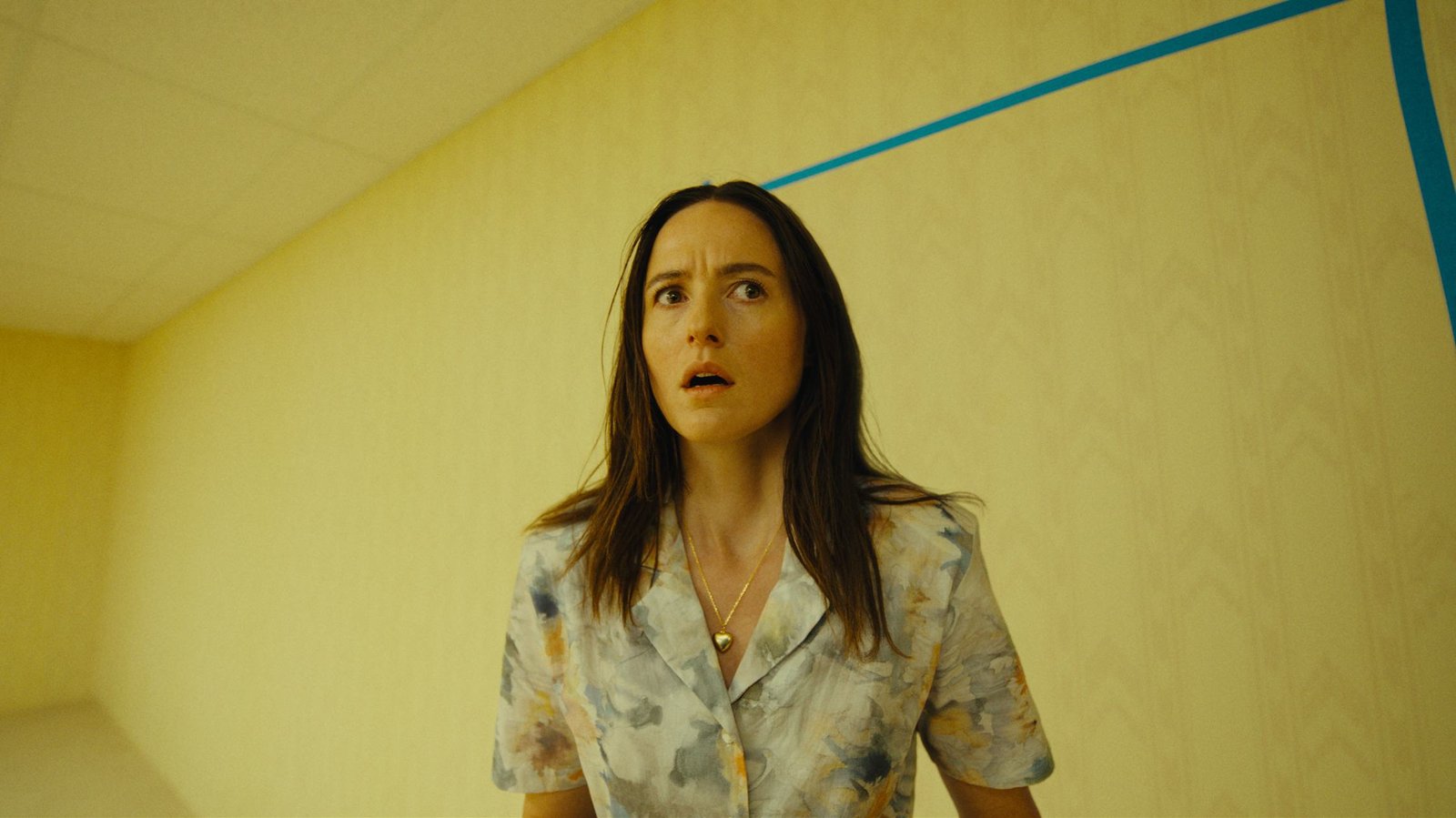 Renate Reinsve as Mary looking up in a yellow liminal Backrooms room, lit by humming fluorescents and marked with blue tape lines